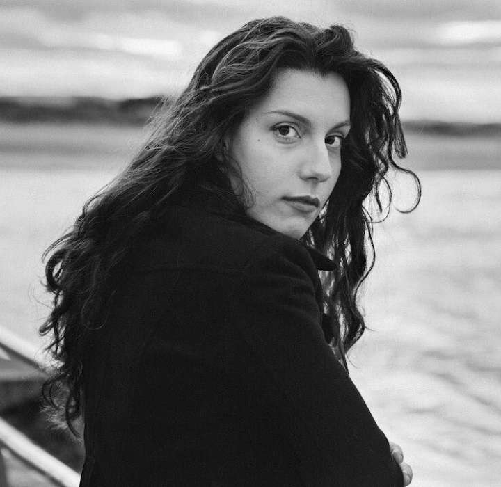 Une femme aux longs cheveux bouclés se retourne, regardant la caméra, avec un paysage côtier flou en arrière-plan.