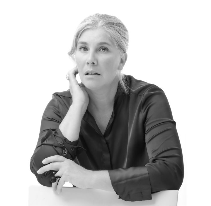 Dieses Bild zeigt Virginie Dhoye, die Gründerin der Nischenparfümmarke ONSKAD, die auf scentissime.com erhältlich ist. Sie ist eine Frau mittleren Alters, die mit ernstem und nachdenklichem Gesichtsausdruck sitzt und direkt in die Kamera blickt. Ihr graues, zurückgekämmtes Haar betont ihr Gesicht. Ihre Haut ist rein und sie wirkt natürlich, ohne starkes Make-up. Eine Hand ruht unter ihrem Kinn. Der Bildhintergrund ist komplett weiß, was einen starken Kontrast zu ihrer Bluse bildet und die Aufmerksamkeit auf ihren Ausdruck und ihre Haltung lenkt. Die gesamte Komposition strahlt eine Atmosphäre von Einfachheit und Tiefe aus und lädt zum Nachdenken ein.