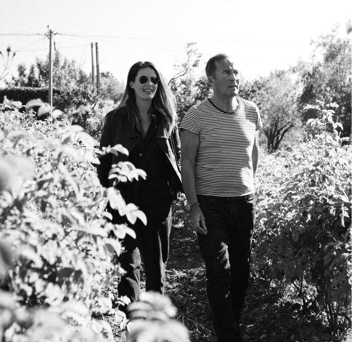 The image shows perfumer Olivier Cresp and his daughter Anaïs walking together in a field or garden. They are surrounded by rows of plants, likely vines or shrubs. The scene is in black and white, creating a nostalgic atmosphere. Anaïs is wearing sunglasses and a leather jacket. Her hair is long and loose. Olivier Cresp is wearing a striped T-shirt and appears relaxed. They are walking side by side, smiling slightly, suggesting a pleasant conversation. The background shows trees and soft light, indicating a sunny day.