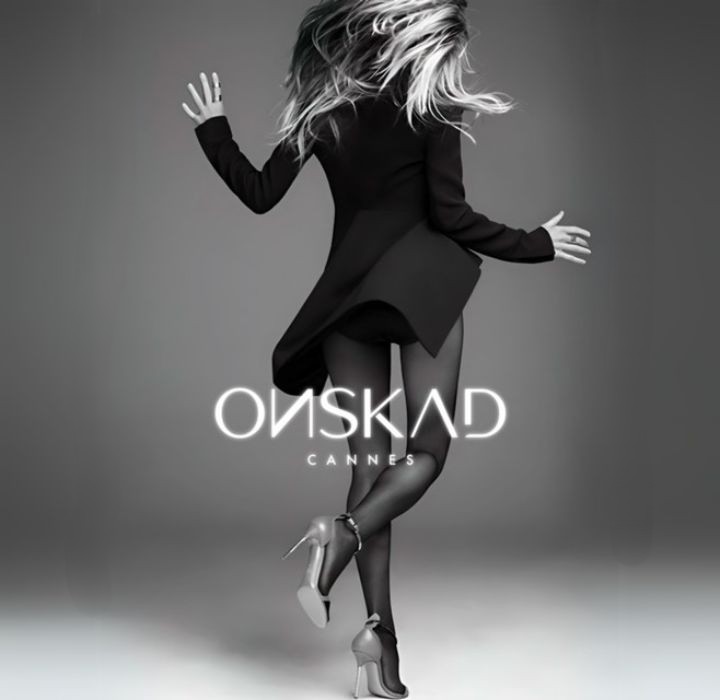 Dieses Bild zeigt eine weibliche Silhouette von hinten, gekleidet in ein elegantes Ensemble. Die Frau trägt einen leicht fließenden schwarzen Mantel, der dynamische Bewegung suggeriert. Ihre Beine sind sichtbar, betont durch schwarze Strumpfhosen und Stilettos, die ihr einen Hauch von Eleganz verleihen.Das lange, leicht gewellte Haar der Frau vermittelt den Eindruck von Bewegung, als würde sie sich anmutig umdrehen. Der Hintergrund ist in Grau gehalten, was die Silhouette betont und eine elegante und moderne Atmosphäre schafft.Am unteren Bildrand steht in weißen Buchstaben das Wort „ONSKAD“, gefolgt von „CANNES“, was auf eine Verbindung zu einem anspruchsvollen Event oder einer Marke hindeutet. Das gesamte Bild strahlt Glamour und Eleganz aus und besticht durch seine Schlichtheit und seinen Stil.