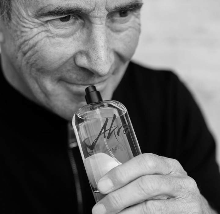 The image shows a portrait of perfumer Olivier Cresp, holding a perfume bottle close to his face. He has a slight smile and a satisfied expression. He wears a black top, which blends in with the blurred and light background, emphasizing his face and the bottle. The perfume bottle is transparent, with the inscription