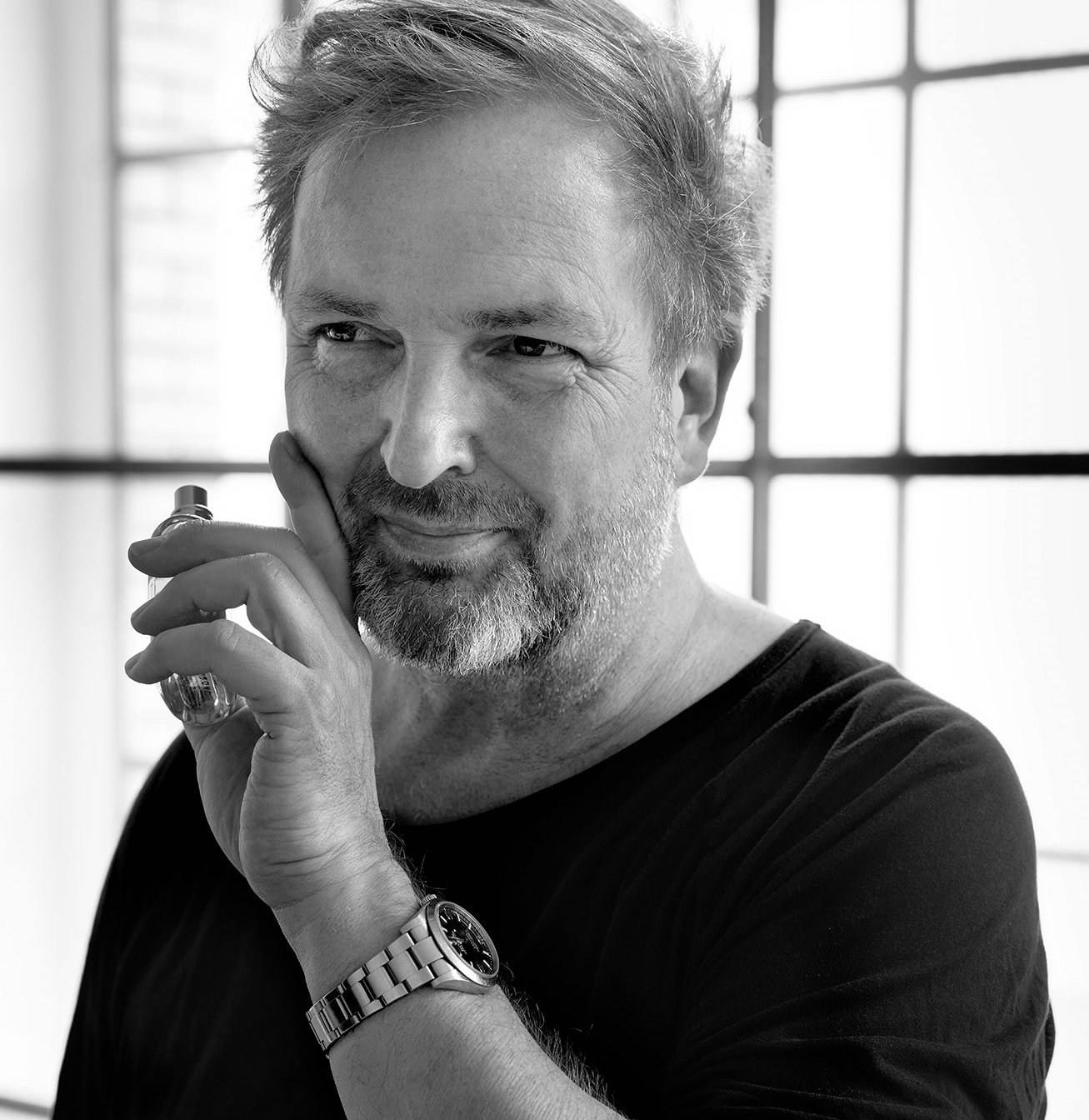 Portrait en noir et blanc de Thomas de Monaco, fondateur de la marque de parfums de niche du même nom. Il tient un flacon de parfum dans sa main gauche, et un a sa main contre le côté de son visage. Il sourit.