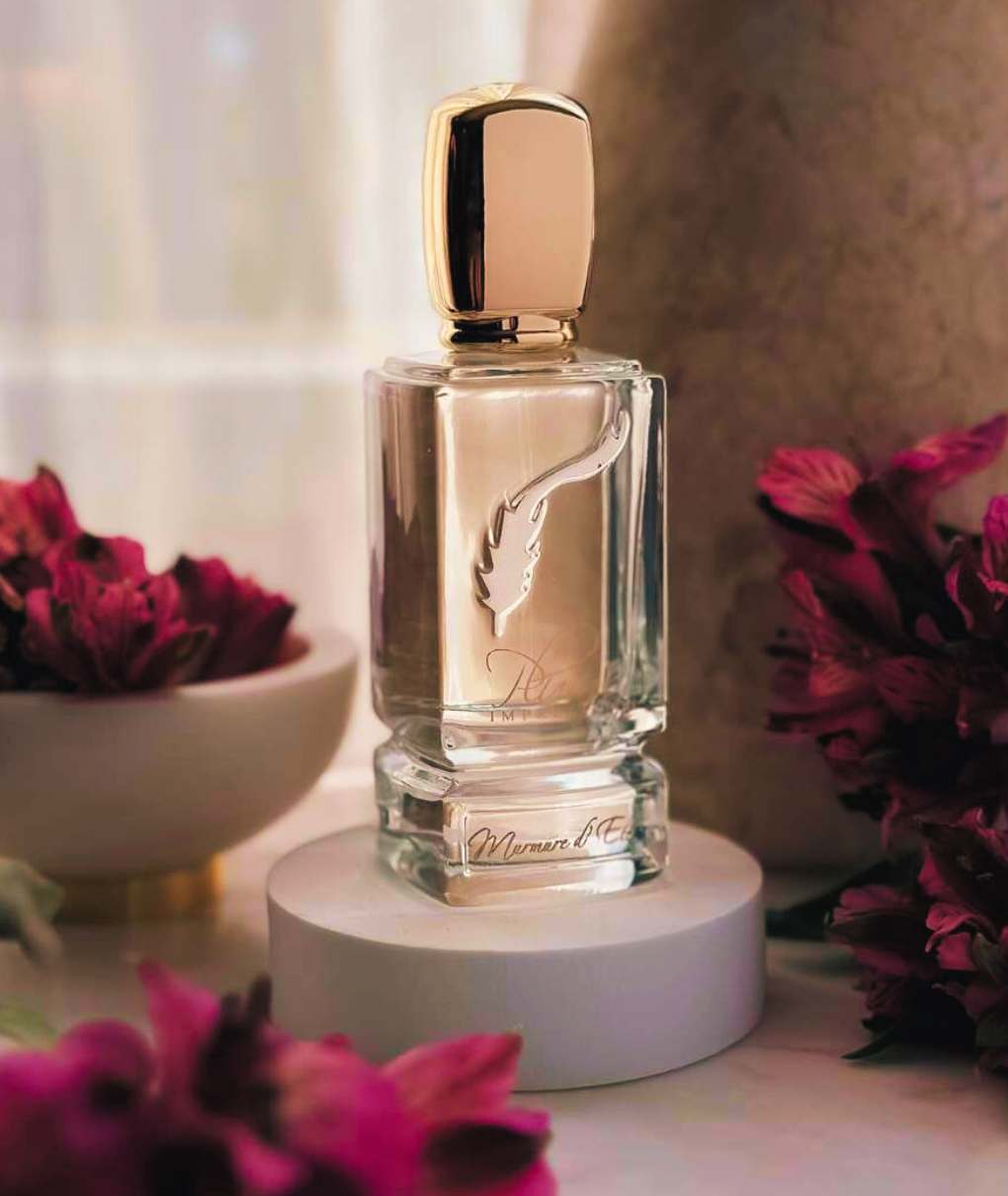 L'immagine mostra un elegante flacone di profumo su una superficie trasparente. Il flacone è trasparente, con una forma rettangolare e un tappo dorato di forma ovale. Il flacone è decorato con un motivo di piume stilizzate, che suggerisce leggerezza e delicatezza.Intorno alla bottiglia ci sono fiori con petali riccamente colorati nelle tonalità del rosa o del viola. Questi fiori sono disposti in modo da incorniciare la bottiglia, creando un effetto estetico armonioso.Sullo sfondo, c'è una leggera sfocatura che suggerisce una texture morbida o un materiale naturale, come la pietra, aggiungendo un tocco organico alla scena. L'insieme evoca un'atmosfera di lusso e bellezza, suggerendo un'esperienza sensoriale raffinata.