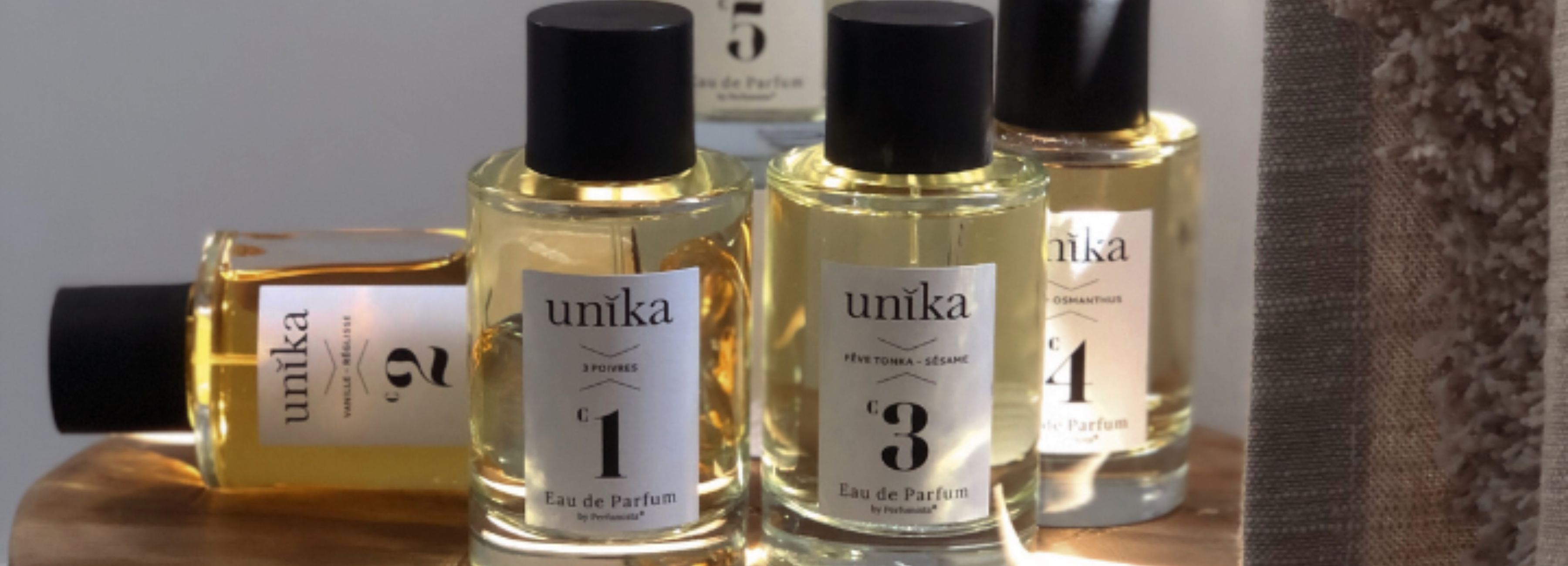 L'image présente plusieurs flacons de parfum de la marque UNIKA, vendue sur la parfumerie en ligne SCENTISSIME. Ils sont élégamment disposés sur une surface en bois. Chaque flacon a une forme similaire, avec un design sobre et sophistiqué. Les étiquettes sont blanches avec le nom