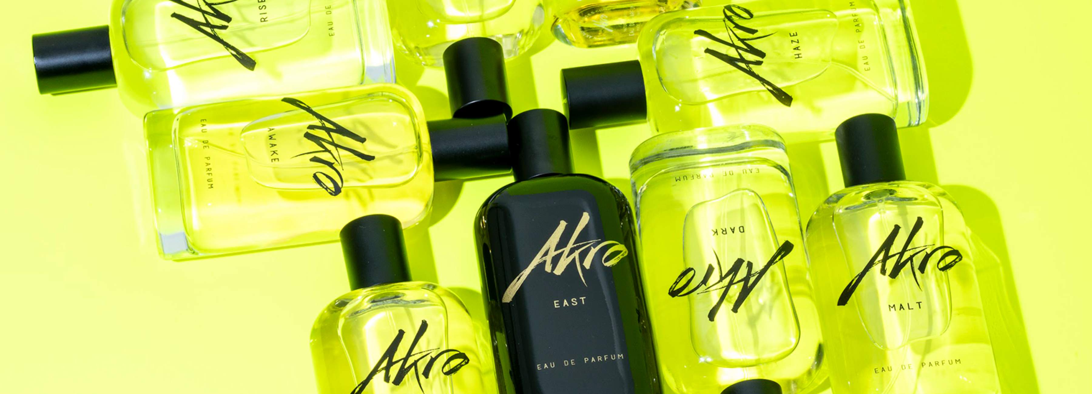 The image shows an arrangement of several perfume bottles from the AKRO FRAGRANCES brand, sold on SCENTISSIME.COM. All lined up against a bright yellow background. Each bottle is rectangular and transparent, allowing a view of the liquid inside, which ranges in color from light to dark yellow. The bottles are labeled with the word