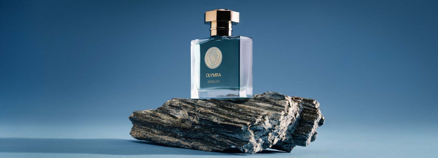 Un flacon de parfum de forme carrée posé sur un rocher, avec un bouchon doré et une étiquette dorée, sur fond bleu.