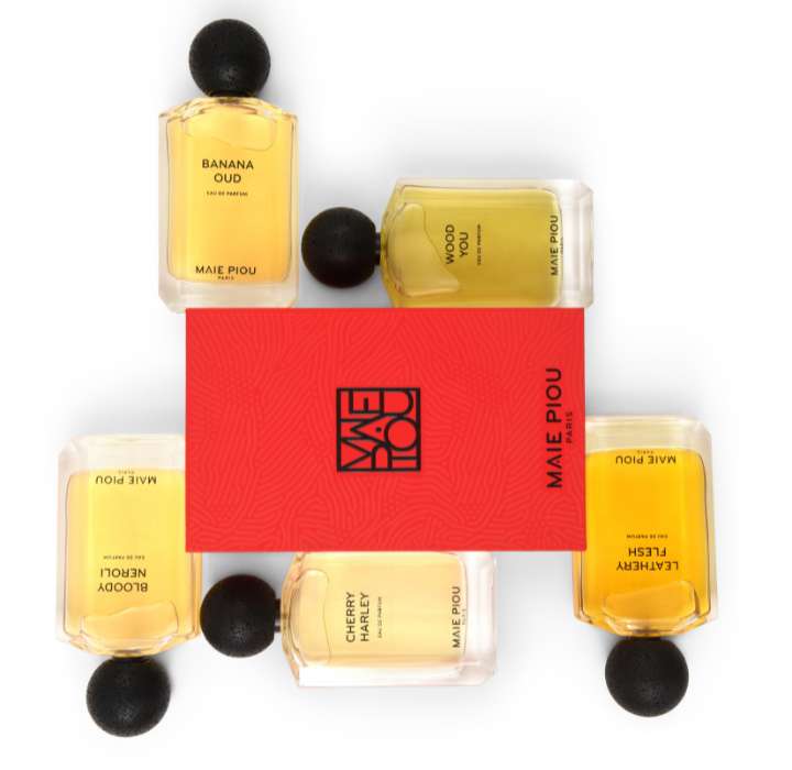 L'image montre un ensemble de flacons de parfum de la marque MAIE PIOU disposés en étoile autour d'une boîte rouge. Chaque flacon a un bouchon noir rond et contient un liquide de couleur différente, allant du jaune clair au doré. Les étiquettes des flacons portent les noms des parfums, tels que