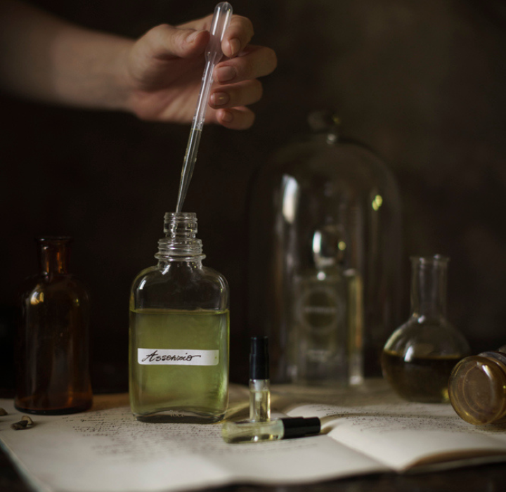 Une scène qui se passe dans un ancien laboratoire de parfumerie. On voit une main qui tient une pipette plongée dans un flacon posé sur un livre ouvert, qui contient un liquide vert