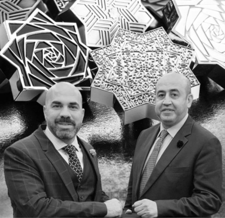 Cette image montre une photo en noir et blanc de Moe Khalaf et Ibrahim Faris , tous deux fondateurs de la marque de parfum de niche KAJAL PERFUMES PARIS, vendue sur le site de vente en ligne SCENTISSIME. Ils se serrent la main. En arrière-plan, une grande photo montre les bouchons des parfums KAJAL.