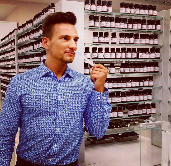 The image shows perfumer Gabriel Gabor. He is standing in a perfume formulation laboratory. He's wearing a blue-patterned shirt and has a pensive look on his face, holding a scent pad in his hand, as if evaluating a fragrance. In the background, we can see shelves filled with bottles of perfume, probably of different essences, neatly organized and labeled. The mood of the image evokes an atmosphere of creativity and discovery in the perfume industry.