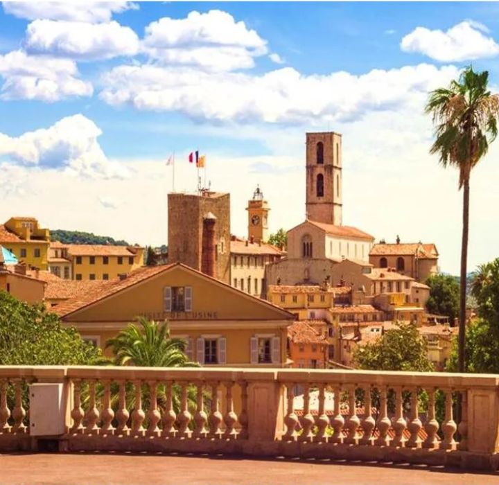 The image features a picturesque cityscape, highlighting the town of Grasse in Provence, with historic buildings. In the foreground, a stone balustrade offers a view of the landscape. The buildings are warmly colored, with tiled roofs and beige and ochre facades. In the background, a church stands out with its high steeple and architectural details. Castle towers can also be seen, adding a medieval touch to the scene. The sky is blue with a few white clouds, creating a pleasant, sunny atmosphere. The overall impression is one of tranquility and historic charm.
