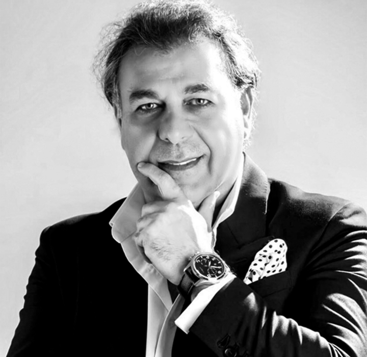 The image is in black and white and shows a smiling man, wearing a dark suit with a light shirt. This is J. Abban, founder of the luxury perfume brand ALEXANDRE J, sold on the online store scentissime.com. He stands with one hand on his chin, and he wears a stylish watch on his wrist. A patterned pocket square is visible in the pocket of his jacket, adding a touch of style. The background is neutral, emphasizing his expression and posture.
