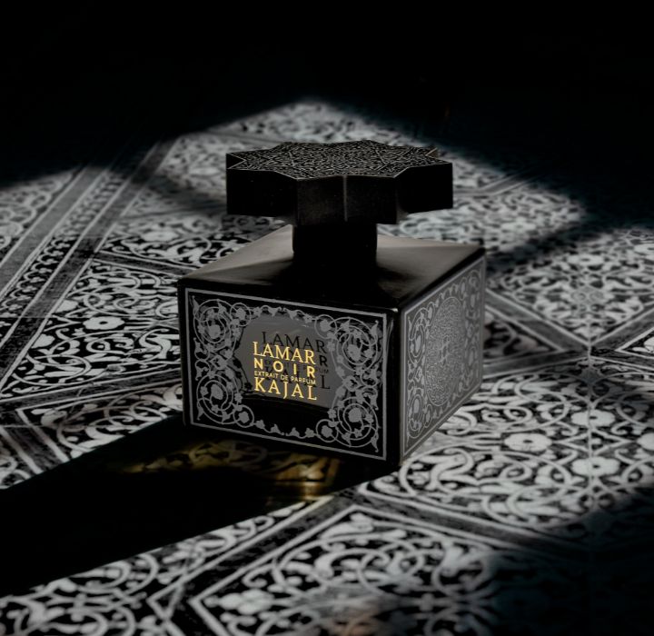 Cette image montre un flacon de LAMAR NOIR, un parfum créé par Kajal. Le design du flacon est complexe et noir, avec une surface réfléchissante qui ajoute à son aspect luxueux. Le nom du produit, « LAMAR NOIR », est affiché en évidence sur le devant du flacon en lettres dorées. L'image se détache sur un fond à motifs arabisants noir et blanc, qui souligne l'élégance et la sophistication du produit. Ce parfum est vendu sur SCENTISSIME