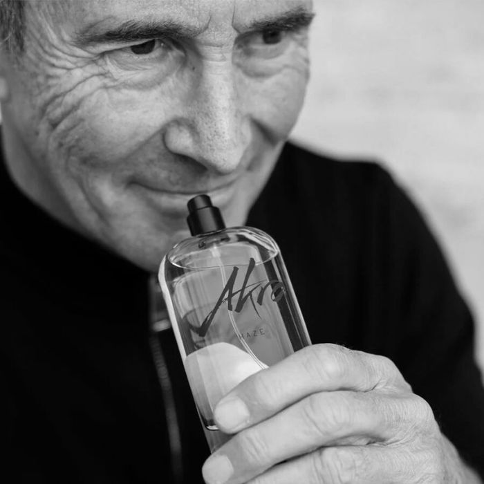 L'image montre un portrait du parfumeur Olivier Cresp, tenant un flacon de parfum près de son visage. Il a un sourire léger et une expression de satisfaction. Son visage est marqué par le temps, avec des rides qui témoignent de son expérience.Il porte un haut noir, qui se fond avec l'arrière-plan flou et clair, ce qui met l'accent sur son visage et le flacon. Le flacon de parfum est transparent, avec l'inscription