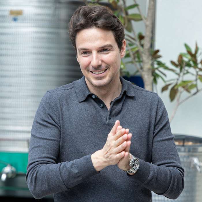A smiling man with brown, slightly curly hair, wearing a gray collared sweater. He stands with hands clasped, as if preparing to speak, in the professional environment of a perfume creation laboratory.