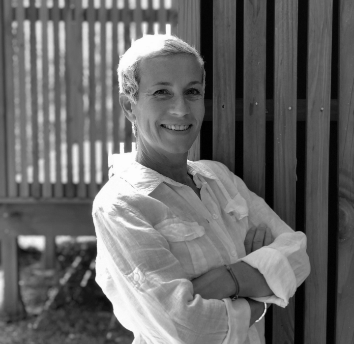 L'image en noir et blanc est une photo de Véronique Stambouli, la parfumeuse qui a créé les parfums de niche UNIKA, vendus sur la parfumerie en ligne SCENTISSIME. Elle est souriante, debout avec les bras croisés. Elle porte une chemise claire, qui contraste avec le fond plus sombre, accentuant son expression amicale et confiante. En arrière-plan, on distingue des éléments en bois et des murs en pierre, créant une ambiance chaleureuse et naturelle. Des plantes ajoutent une touche de verdure.