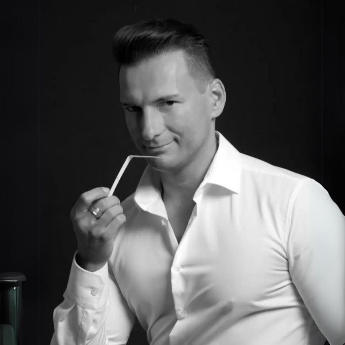 The image shows Gabriel Gabor in portrait form, dressed in a well-fitting white shirt. He has a confident expression, with a slight smile, and is holding a sniffing dab between his fingers, as if evaluating a fragrance. The background of the photo is black, highlighting his face and outfit. His hair is neatly styled, adding to his elegant allure. The lighting is soft, creating a contrast that accentuates his features and the professional atmosphere of the scene.
