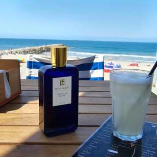 The Atlantic was turning gold when I sat down at the bar.

I had been walking along the Portuguese coast since late morning, with the sea beside me and Infinite Blue on my skin. Hours had passed since I had sprayed it on, yet every movement brought something back. A trace of bergamot. The cool, mineral impression of the ocean. Lavender and rosemary warmed by the afternoon sun.

The bottle was sitting on the table in front of me.

Infinite Blue.

I looked at it for a moment, then out towards the horizon. From where I was sitting, the two seemed almost connected. The bottle on the table, the endless blue beyond it.

I had come to Portugal to slow down. As the owner of Scentissime, my days are usually filled with names, launches, samples, messages, deliveries, numbers and decisions. Fragrance has become my profession, but I still need places where I can experience it without thinking about business.

This was one of them.

A glass arrived. The bar was quiet, the kind of place where nobody seems to be in a hurry. In front of me, the Atlantic was moving slowly beneath the evening light.

I raised my wrist.

Infinite Blue had changed.

The freshness of the opening had disappeared hours ago. What remained was warmer, softer, more intimate. The aromatic character of rosemary and lavender was still there, but now surrounded by woods, amber and the gentle sweetness of tonka bean. Patchouli gave it a darker undertone.

It smelled of the sea remembered at sunset rather than the sea at noon.

That is what I like about wearing a fragrance all day. You stop judging it. You stop analysing its notes. You simply live with it.

A breeze came in from the ocean and lifted the sleeve of my shirt. For a second, the scent moved around me. Marine air, aromatic herbs, woods and amber. Freshness and warmth existing together.

I smiled.

Perhaps this is what I am looking for when I choose a fragrance.

Not something that announces itself. Something that accompanies you.

Something that changes with the hours, with the weather, with the place, and eventually with your own mood.